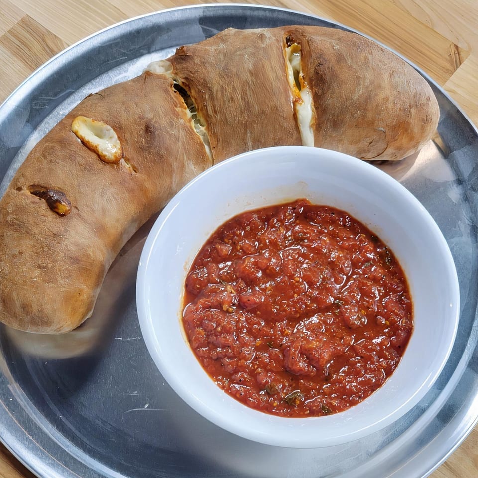 A golden-brown calzone with melted cheese peeking through, served with a side of chunky marinara sauce.