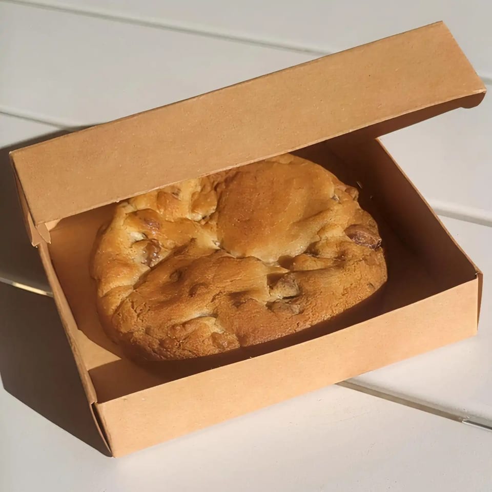 A large, warm chocolate chip cookie sits inside an open brown takeout box on a white surface.