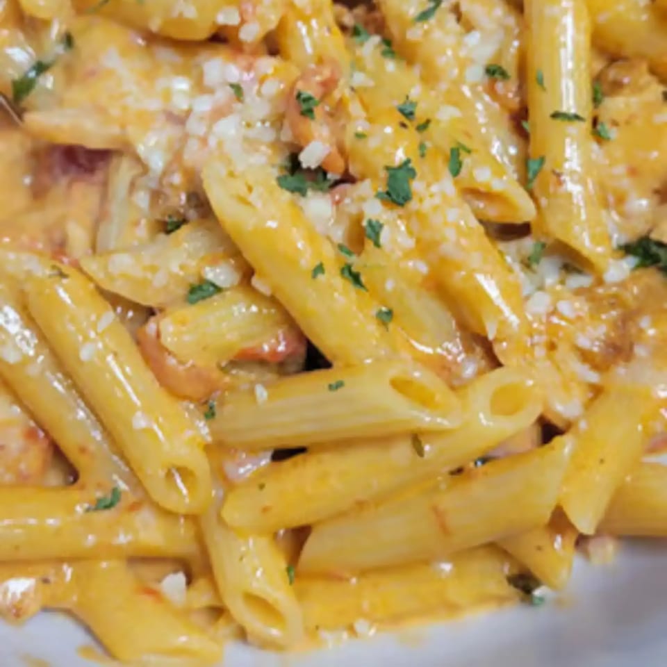 Close-up of penne pasta in a creamy orange sauce, topped with white cheese and green herbs.